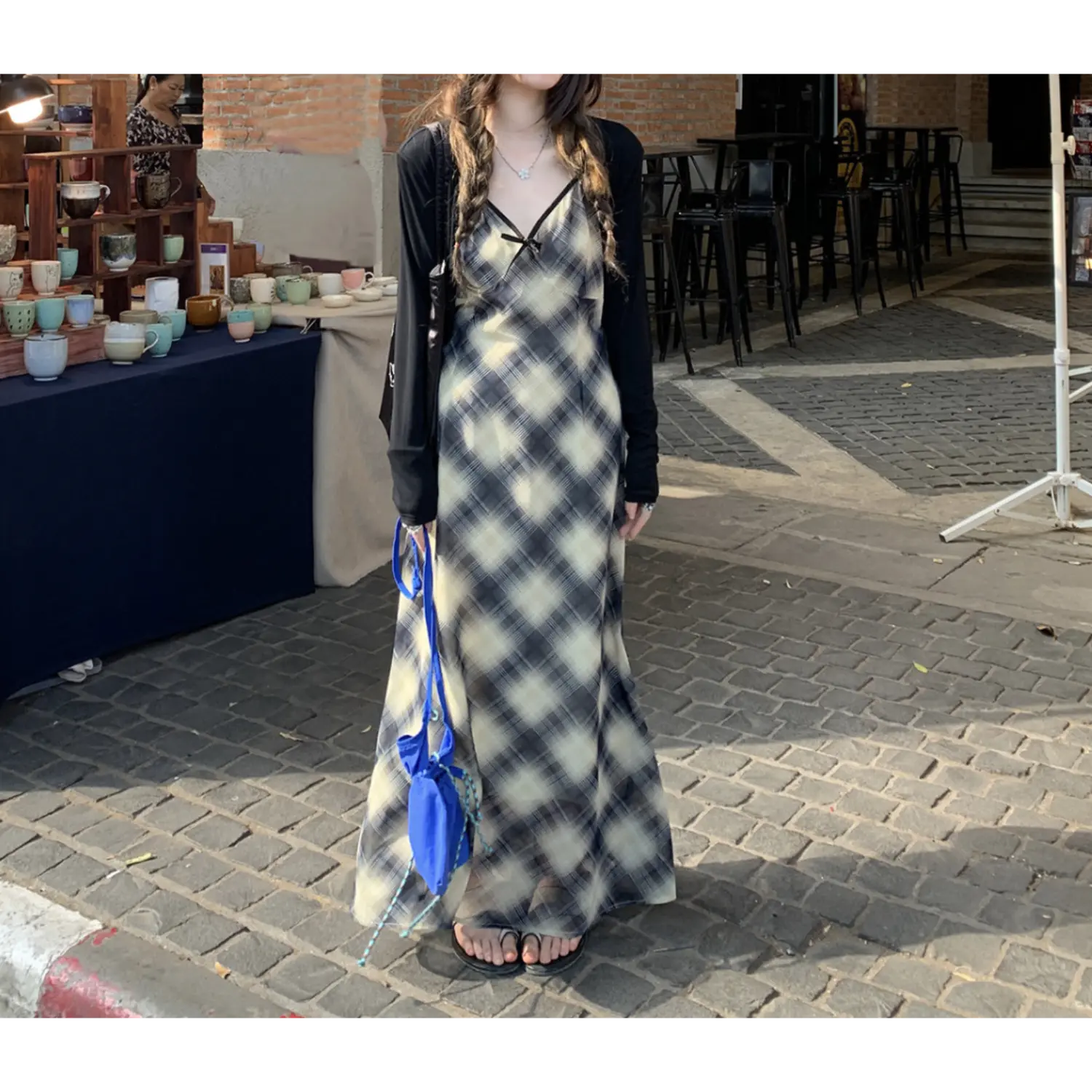 Casual outdoor market lifestyle shot of sheer chiffon plaid slip maxi dress, diagonal cream black check pattern, thin spaghetti straps, flowy floor-grazing skirt, model paired with black slide sandals, bright blue drawstring crossbody bag, vintage brick street & ceramic stall backdrop, natural daylight