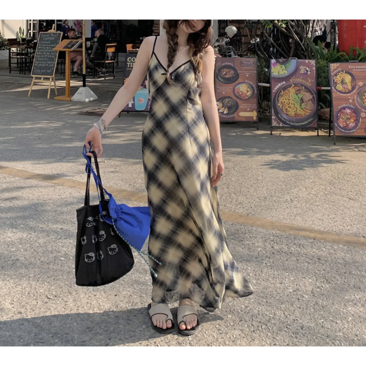 Relaxed roadside lifestyle shot of bias plaid chiffon maxi slip dress, faded cream black diagonal grid print, thin adjustable cami straps, flared long skirt, model holding double tote bags, street food menu sign & tropical greenery background