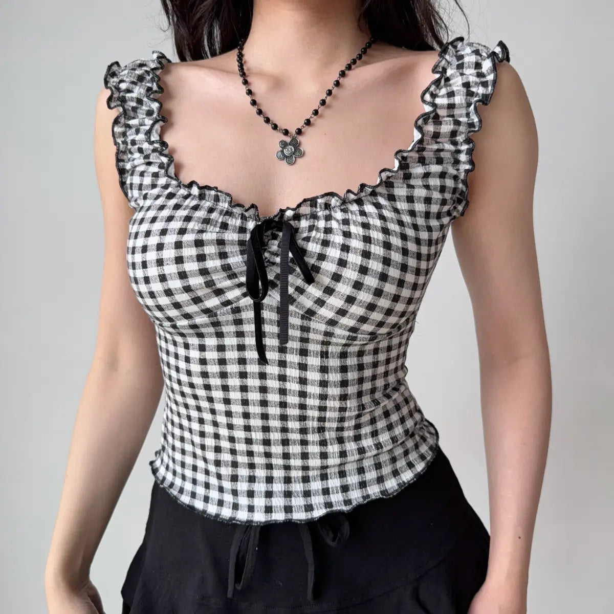 Model wearing black and white gingham cami top with ruffled scallop trim, sweetheart neckline, and front tie detail