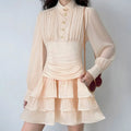 Model wearing blush pink mini dress with pleated collar, gold button placket, sheer long sleeves, ruched waist, and tiered ruffle skirt