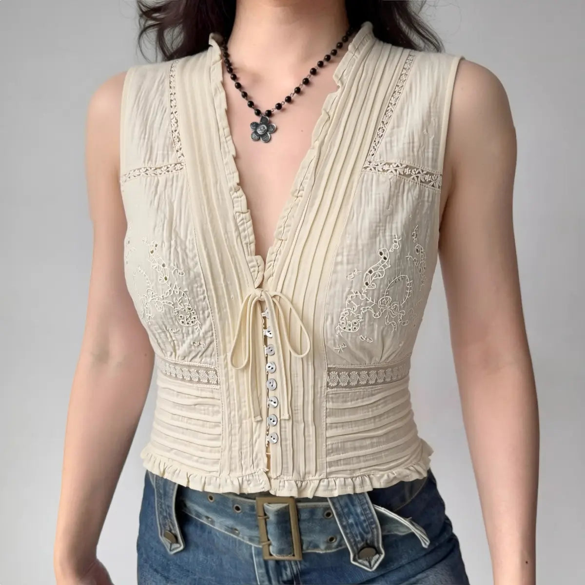 Model wearing cream lace sleeveless vest with deep V-neck, ruffled trim, front drawstring tie, and cropped fitted silhouette