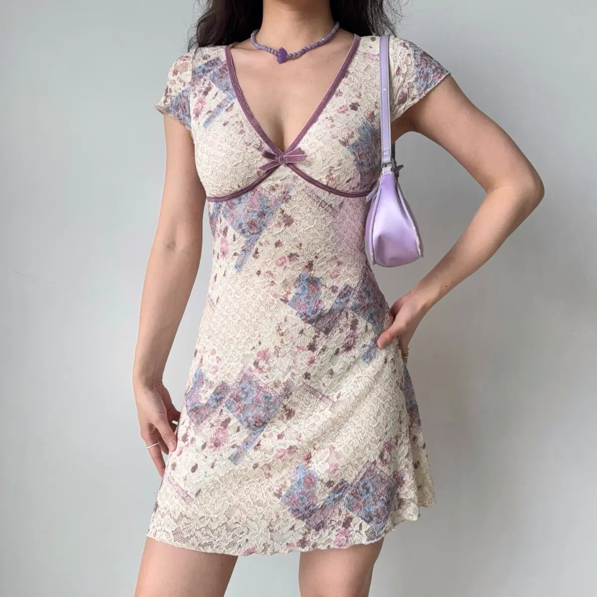 Model wearing cream base lavender floral lace mini dress with short sleeves, deep V-neckline and purple contrast trim
