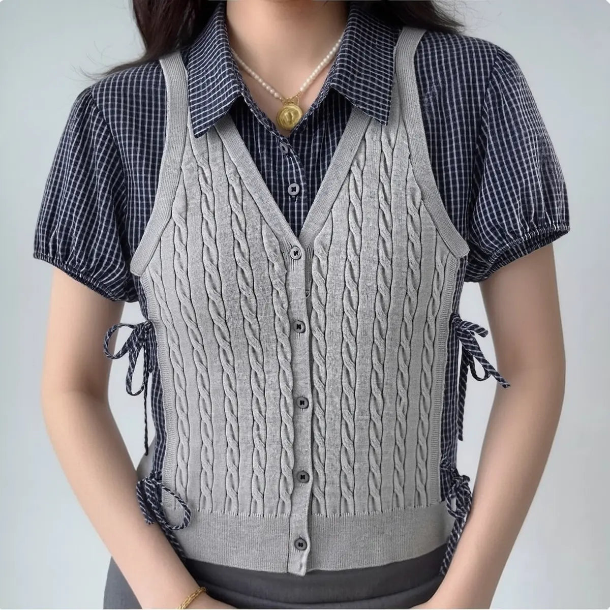 Model wearing grey cable-knit vest over navy gingham shirt with puff sleeves, preppy layered two-piece outfit