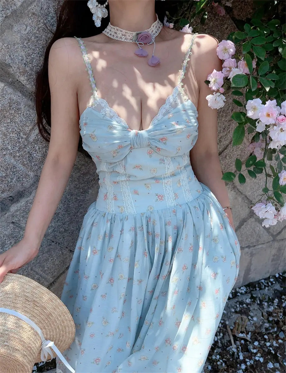 Model wearing light blue floral print cami midi dress with lace-trimmed bust, front bow detail, spaghetti straps, and flowy tiered skirt