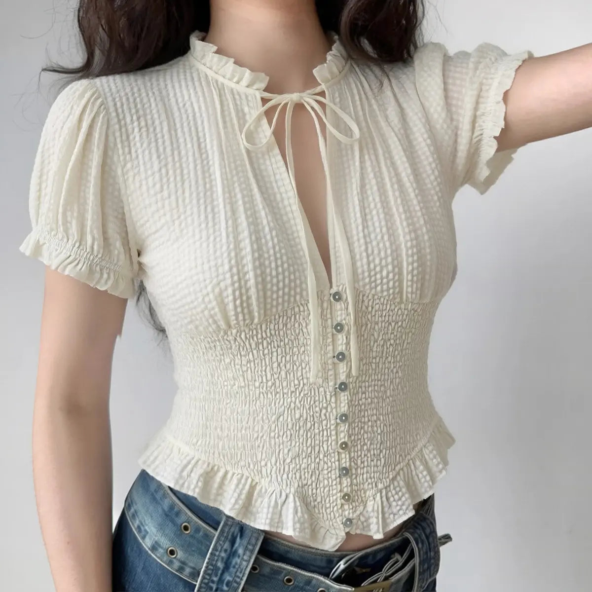 Model wearing off-white textured puff-sleeve blouse with tie neck and smocked waist, vintage-inspired cropped top