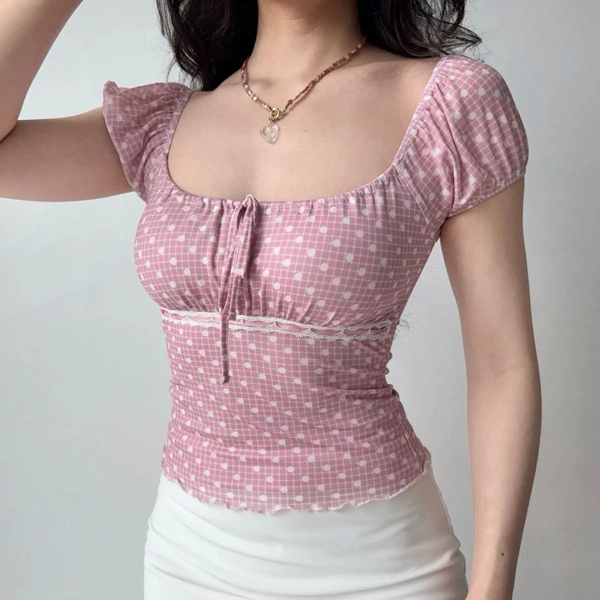 Model wearing pink-white polka-dot top, raising arm to show puff sleeve and fit, with square neckline and lace trim
