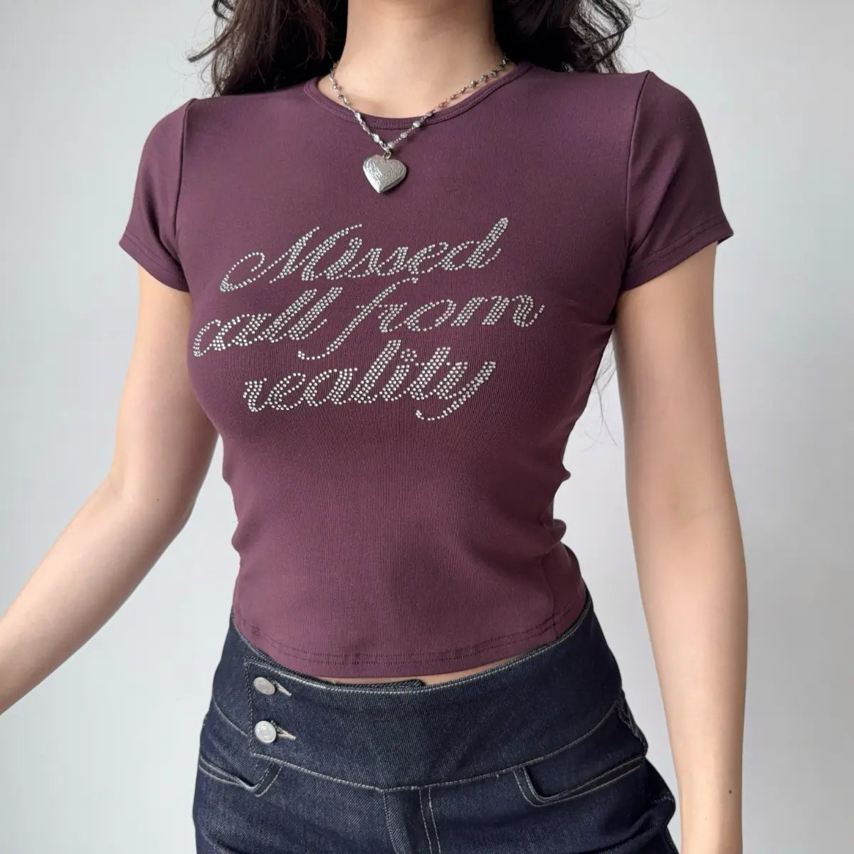 Model wearing plum-colored short-sleeve t-shirt with "Missed call from reality" script print, crew neck, and form-fitting cropped silhouette