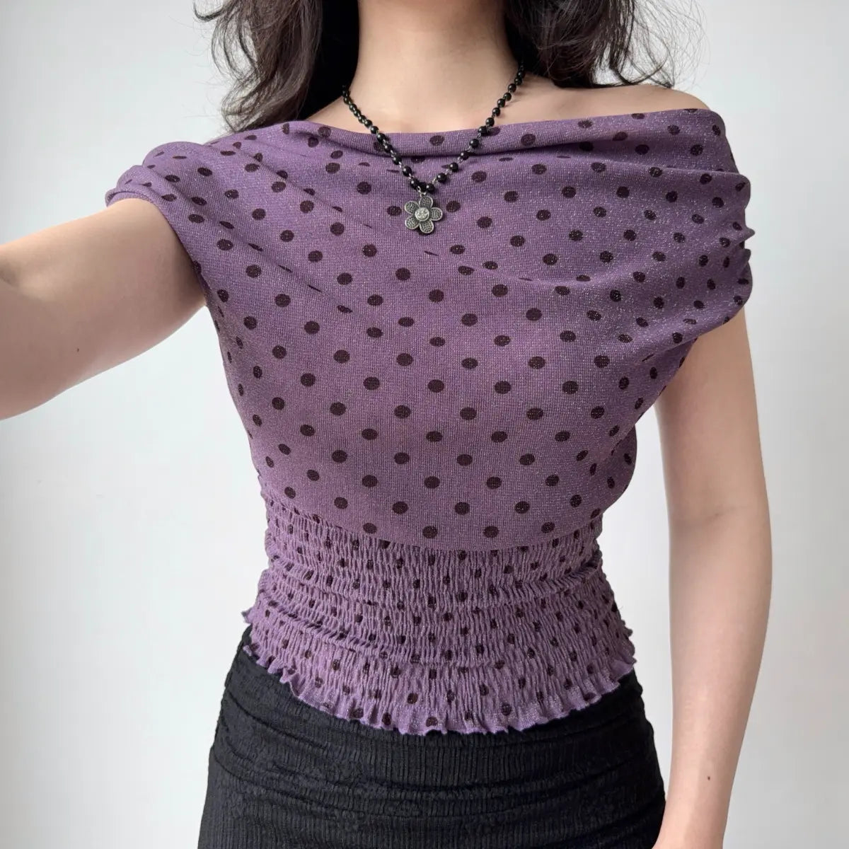 Model wearing purple polka-dot off-the-shoulder top with smocked waist and ruffled hem, raising arm to show fit and fabric