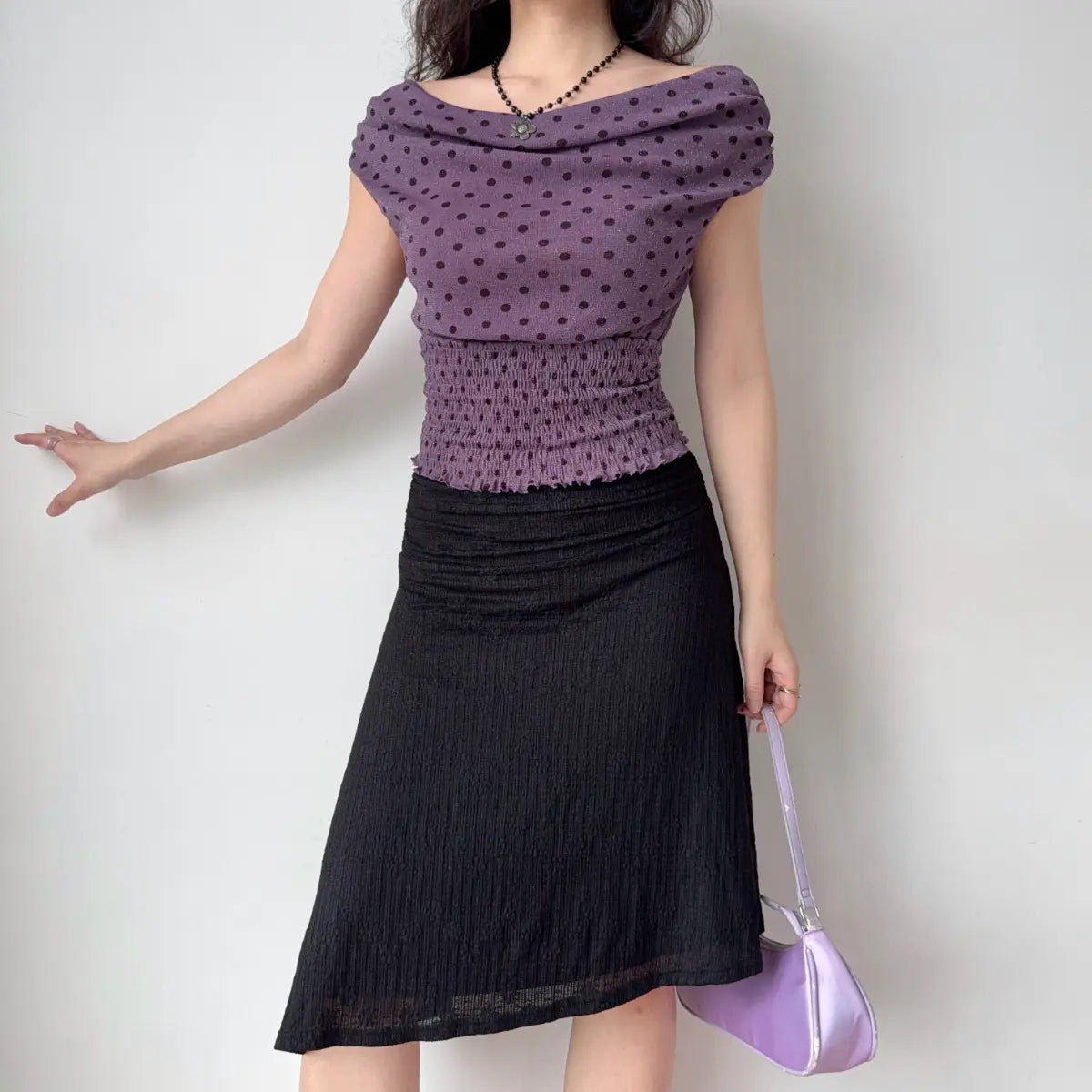 Model wearing purple polka-dot off-the-shoulder top paired with black textured midi skirt, styled with a matching purple bag