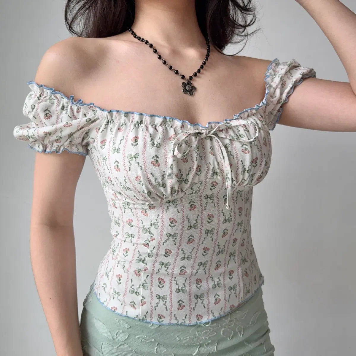 Model wearing white floral-print off-the-shoulder top with blue ruffle trim, puff sleeves and front bow detail, raising arm to show fit

