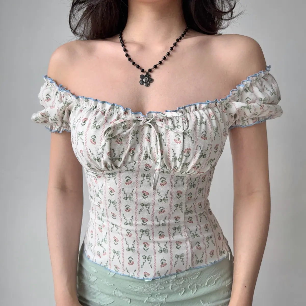 Model wearing white floral-print top styled as off-the-shoulder, with blue ruffle trim, puff sleeves and front bow detail, fitted corset-style blouse