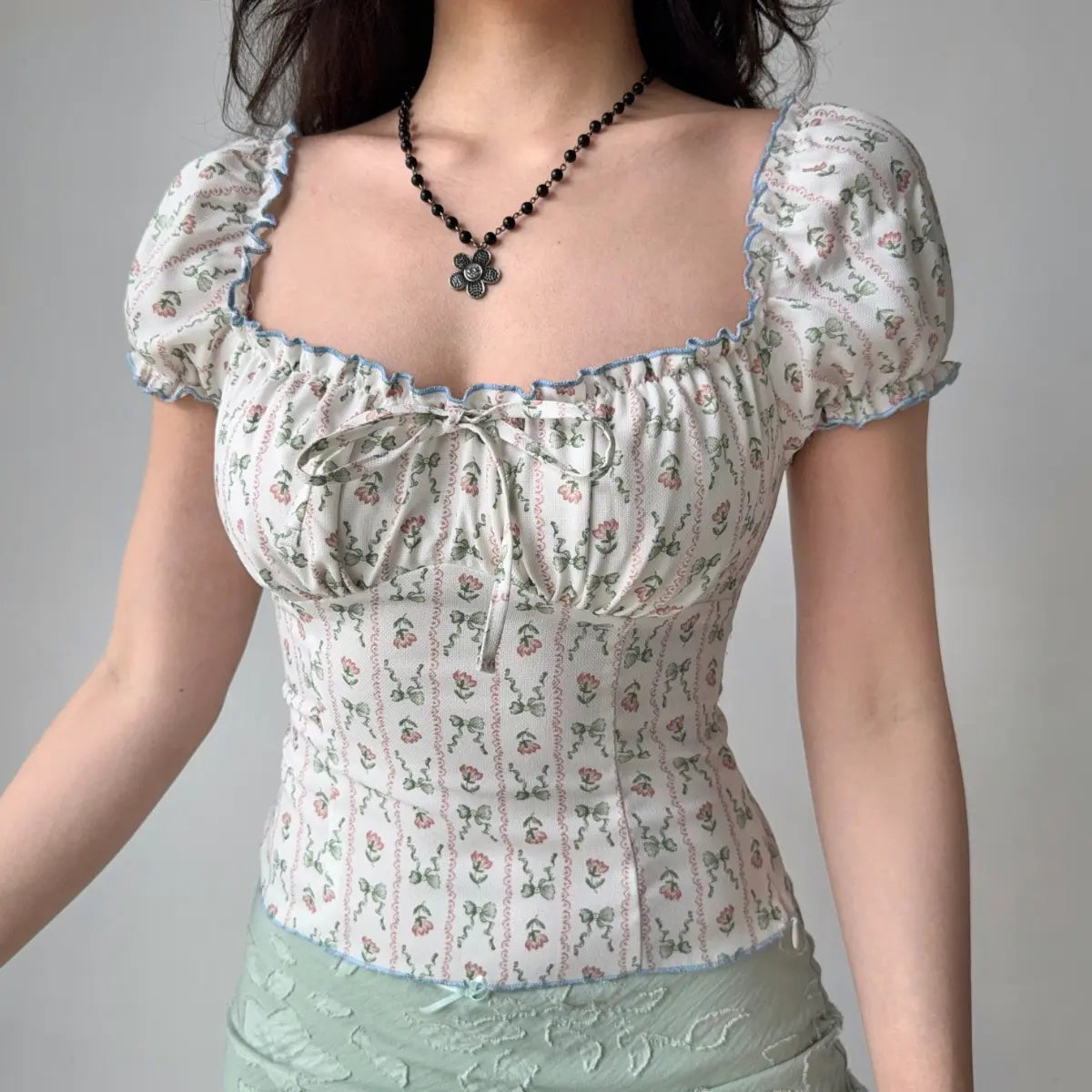 Model wearing white floral-print top with square neckline, blue ruffle trim, puff sleeves and front bow detail, fitted corset-style blouse