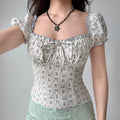 Model wearing white floral-print top with square neckline, raising arm to show puff sleeve and fit, with blue ruffle trim and front bow detail