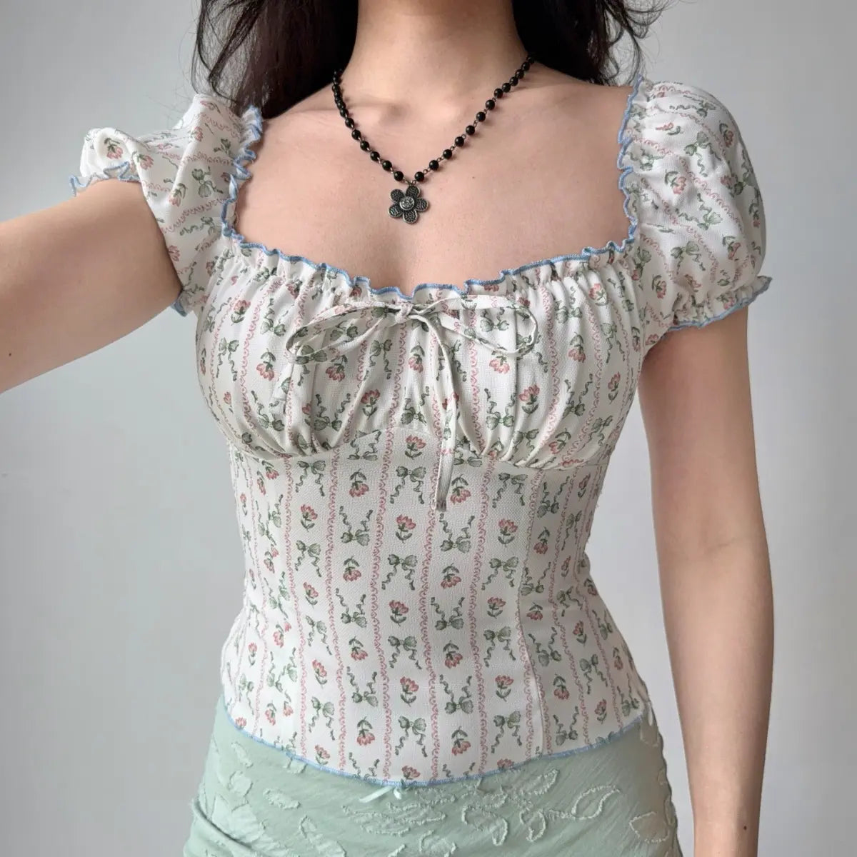 Model wearing white floral-print top with square neckline, raising arm to show puff sleeve and fit, with blue ruffle trim and front bow detail