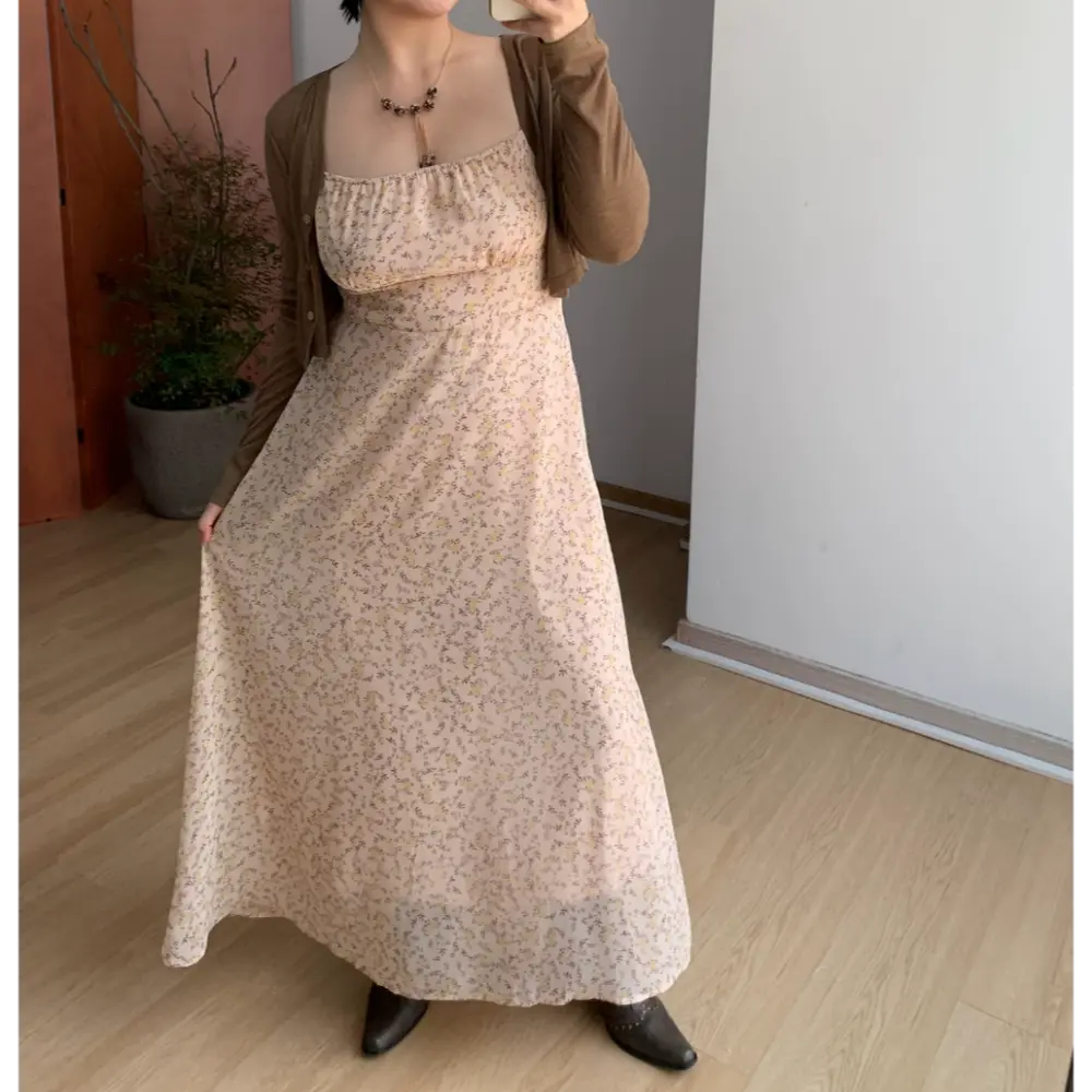 Medium full indoor mirror lifestyle shot of pale apricot micro floral spaghetti strap maxi dress, layered brown cropped cardigan, model gently lifting side skirt hem to show flowy fabric