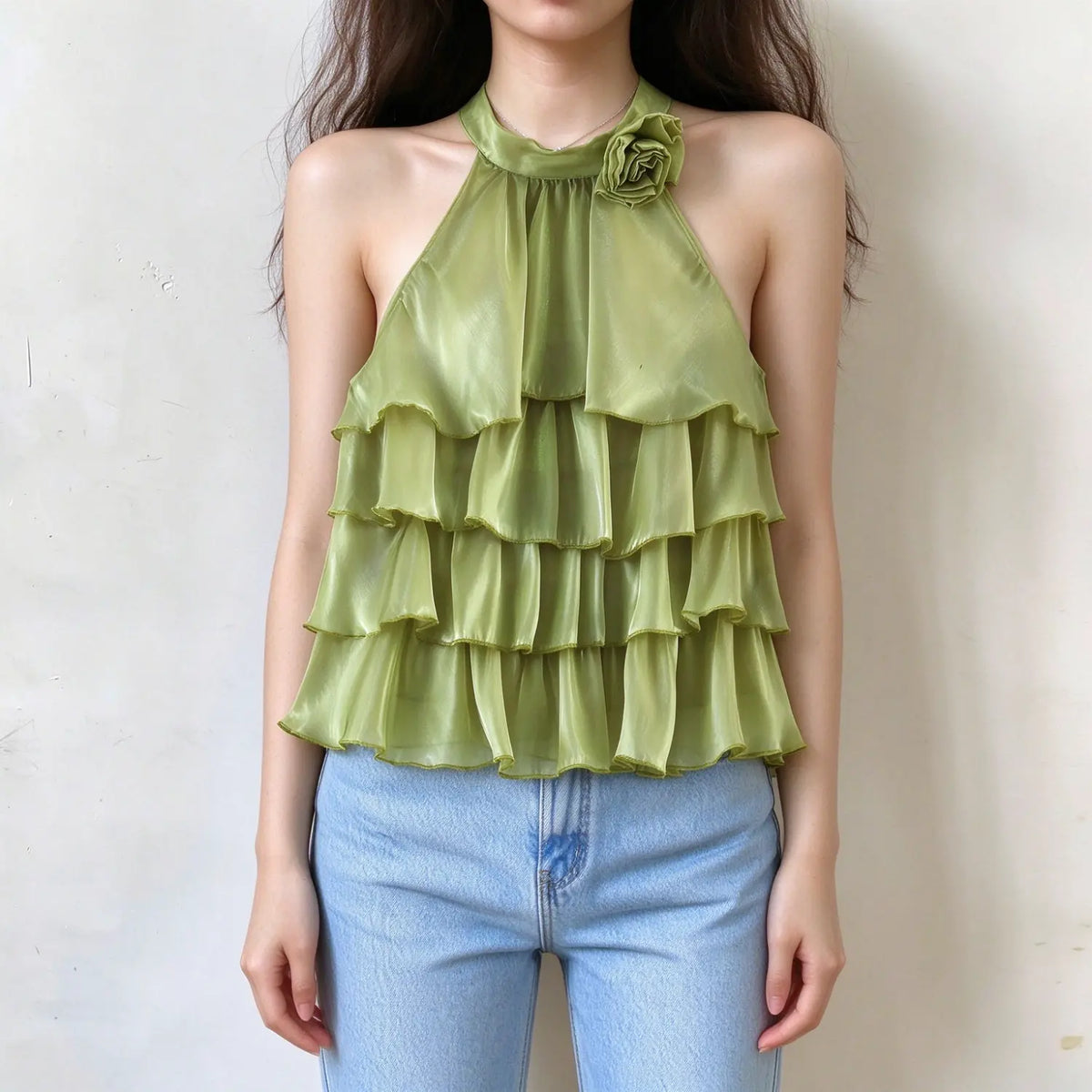 Model wearing avocado green halter neck tiered ruffle top with fabric flower decoration, sheer glossy fabric, sleeveless backless design, paired with light blue jeans, plain white wall background
