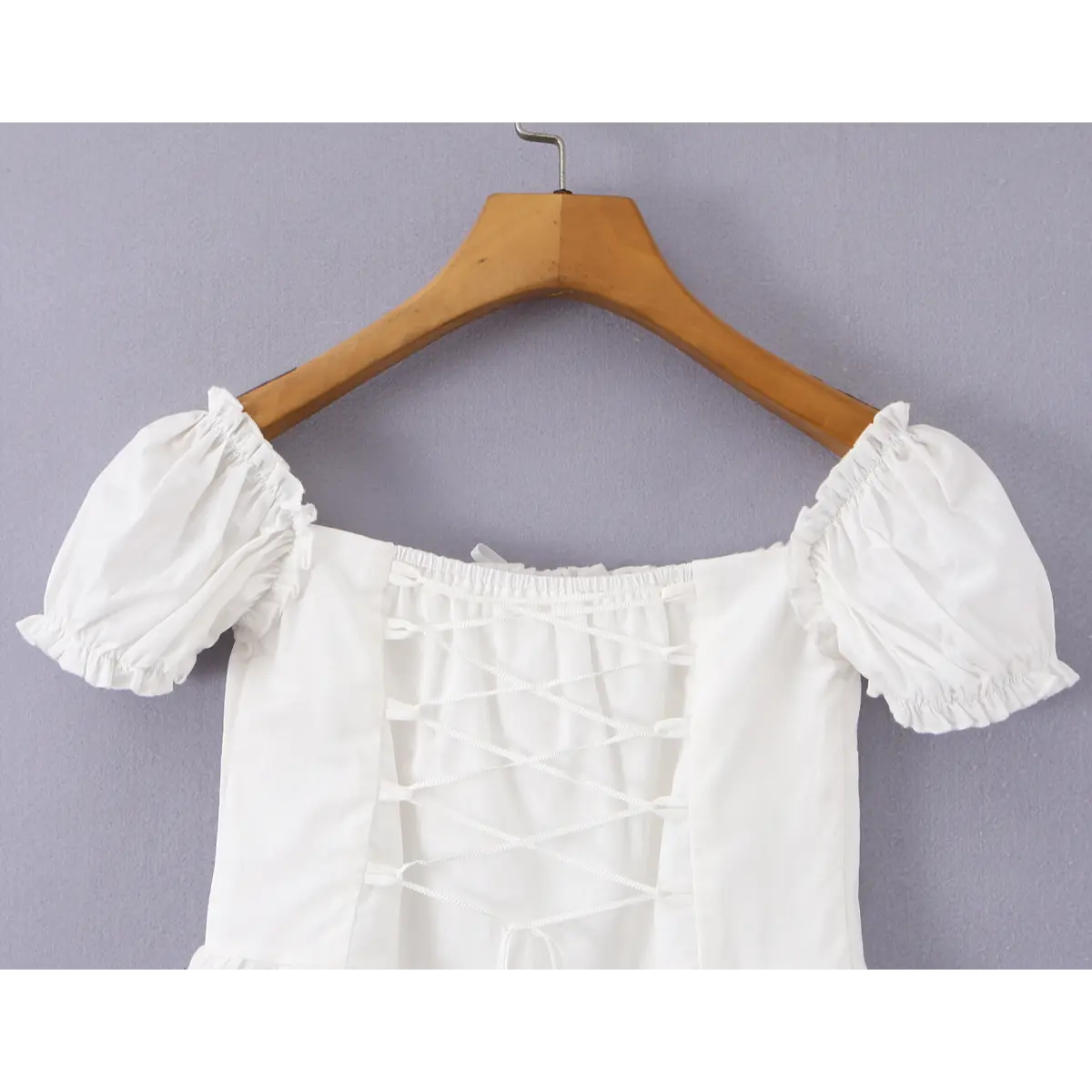 Back view of a white off-shoulder peplum top on a wooden hanger, showcasing the corset-style lace-up closure and puff sleeves.