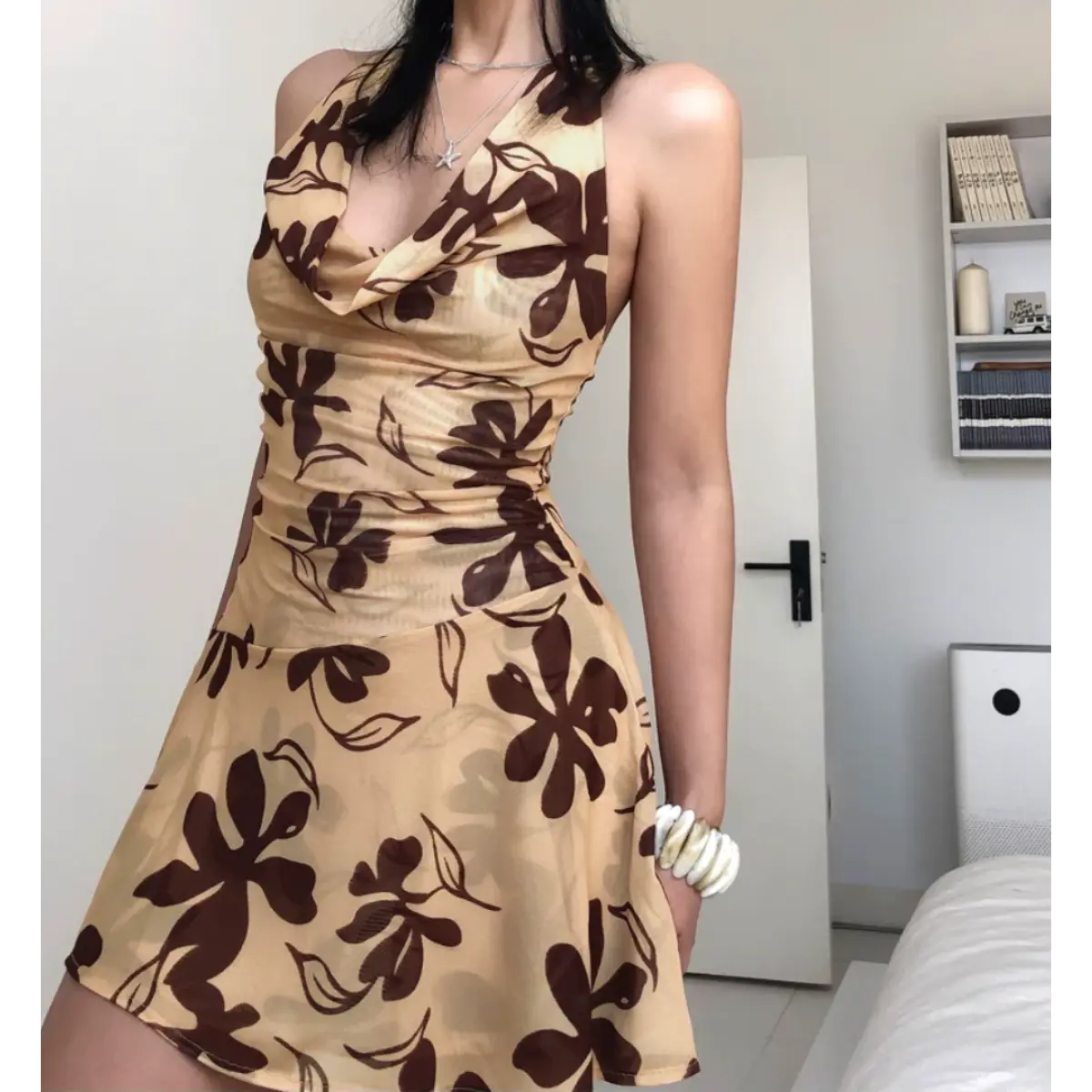 Woman wearing a beige halter mini dress with a brown floral print, featuring a cowl neckline, mesh fabric, and flared skirt.