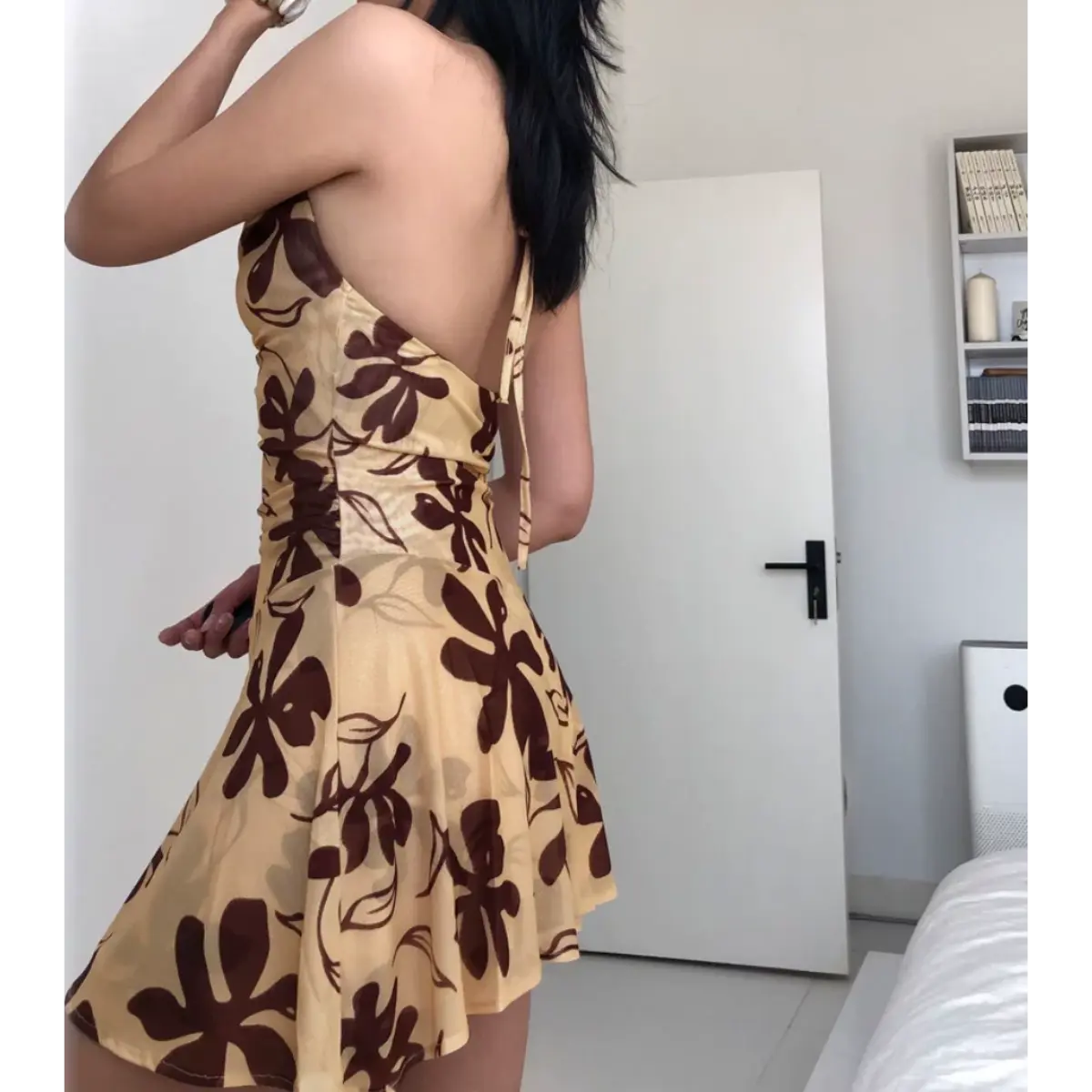 Back view of a woman wearing a beige halter mini dress with a brown floral print, showing the open back and halter tie detail.