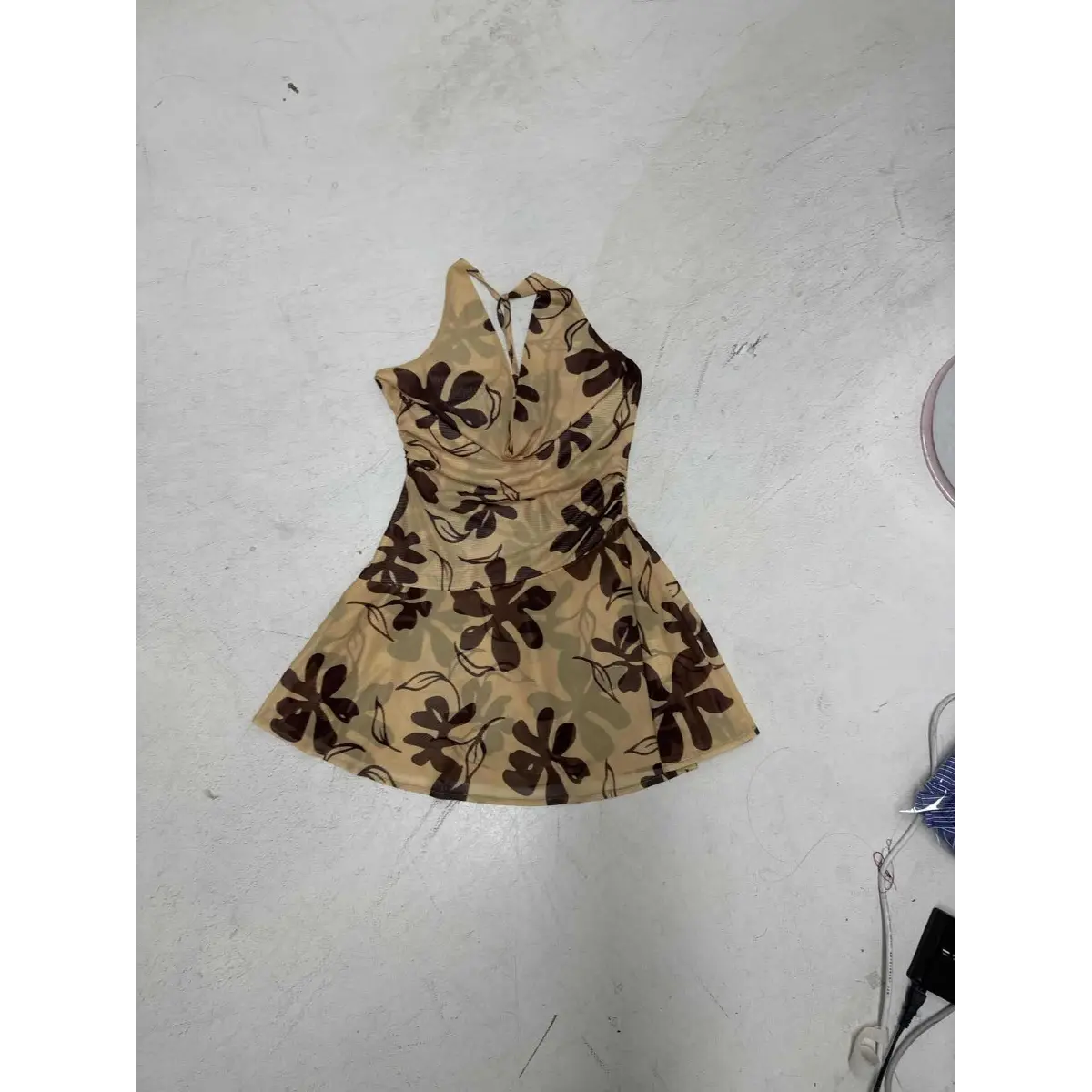 Top-down view of a beige halter mini dress with a brown floral print, featuring a cowl neckline, mesh fabric, and flared skirt, laid flat on a light gray background.