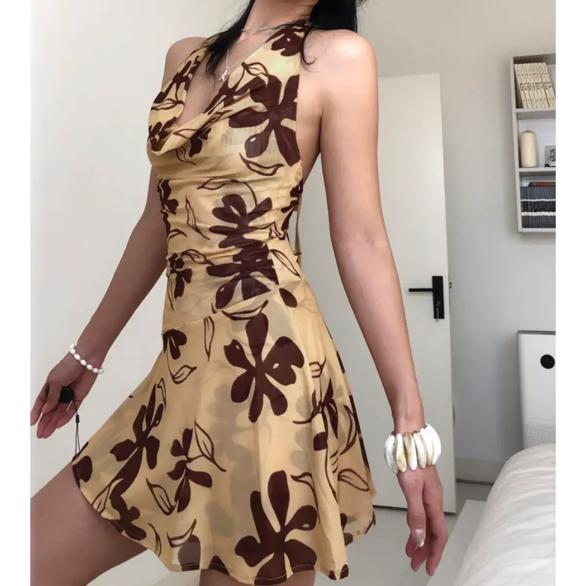 Woman wearing a beige halter mini dress with a brown floral print, posed with arms spread to show the dress's silhouette and open back.