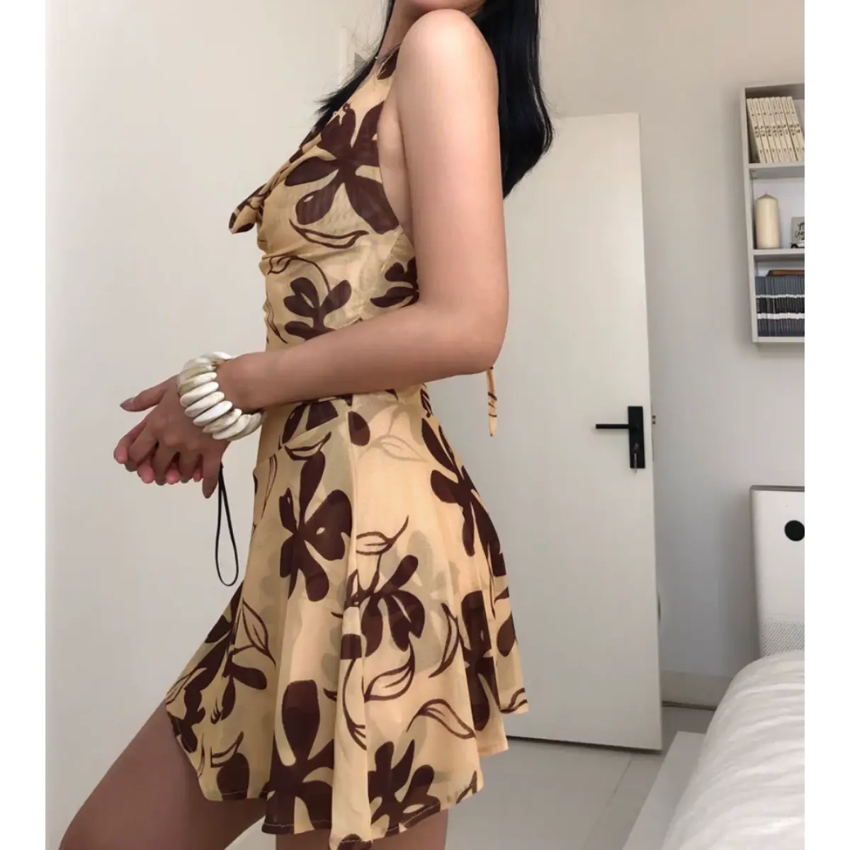 Three-quarter view of a woman wearing a beige halter mini dress with a brown floral print, showing the side profile and flared skirt.