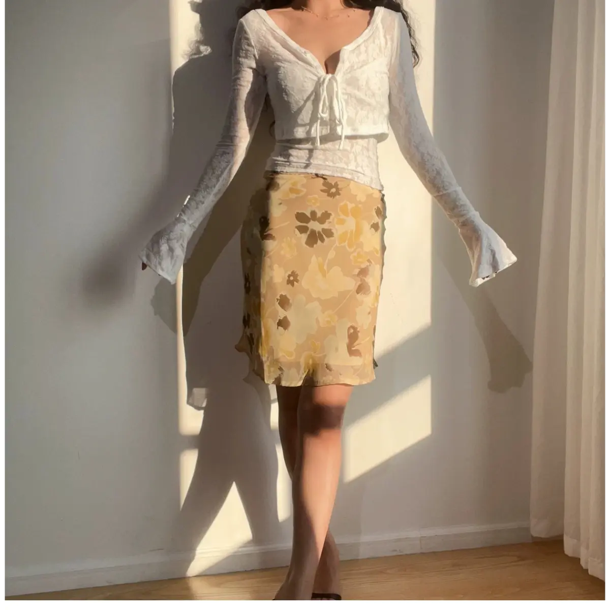 Full-body view of a woman wearing a beige floral chiffon midi skirt with brown and yellow flowers, paired with a white lace long-sleeve cropped top.