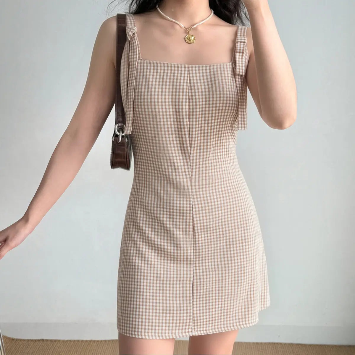 Model wearing a beige gingham mini dress with bow-tied straps, styled with a light blue heart-shaped bag and gold necklace.