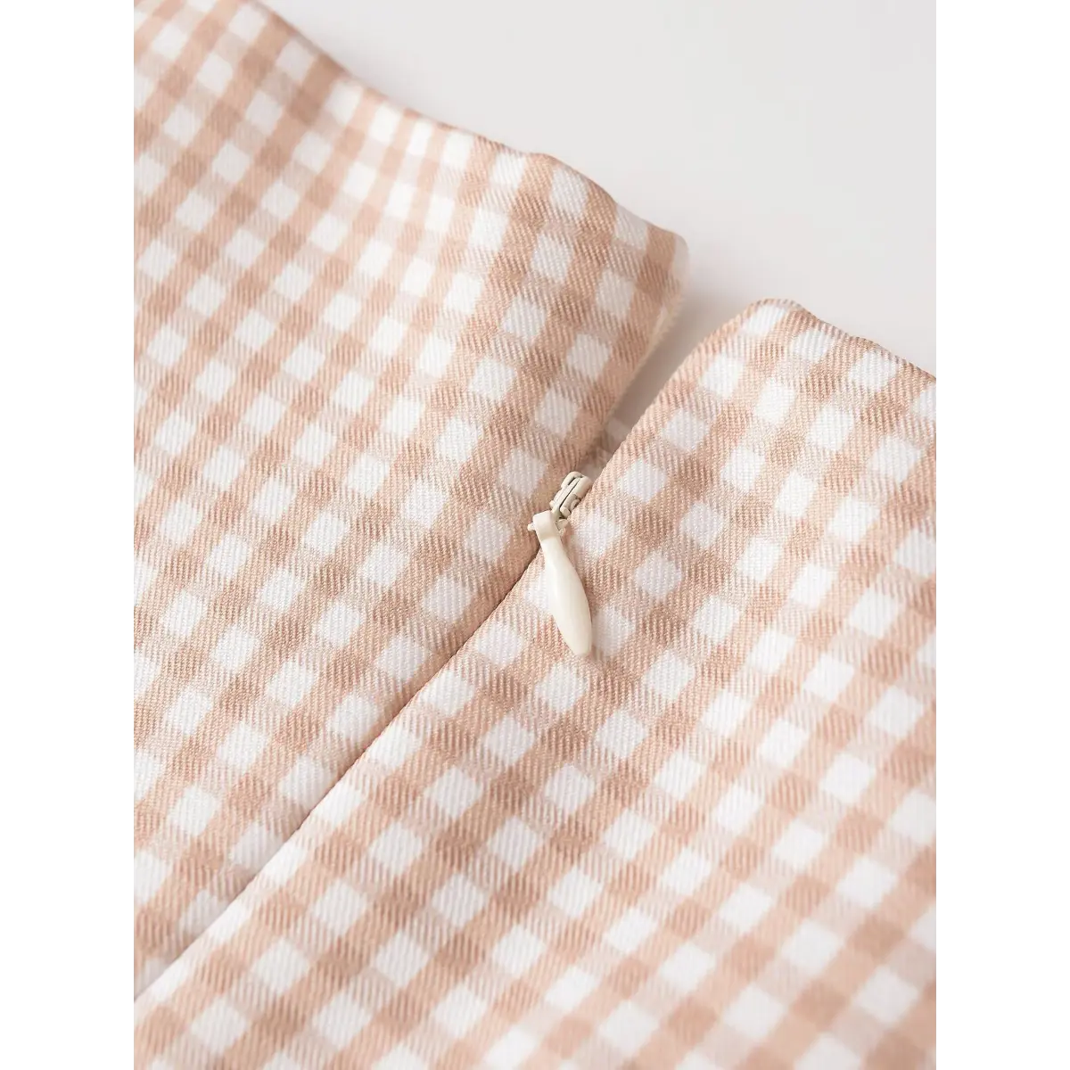 Close-up detail shot of the back zipper on a beige gingham mini dress, showing the zipper pull and gingham pattern.