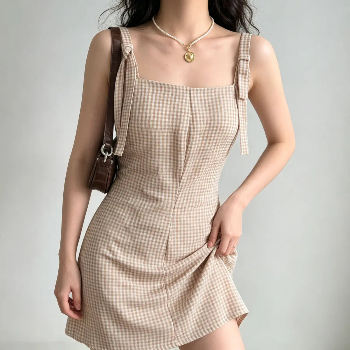 Model wearing a beige gingham mini dress with tie straps, styled with a brown leather bag and pearl necklace, holding the skirt to show the fabric texture.