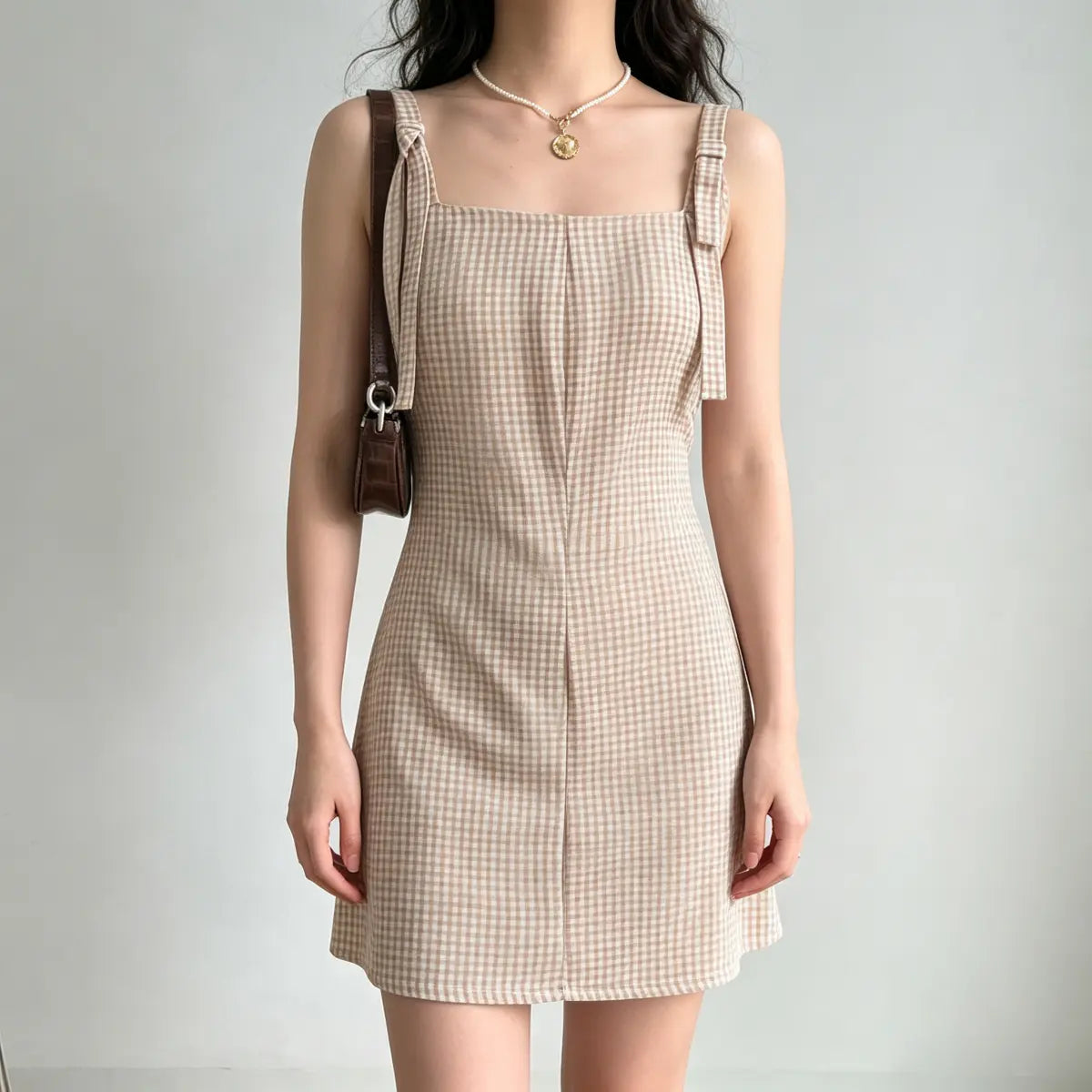 Model wearing a beige gingham mini dress with tie straps, styled with a brown leather bag and pearl necklace, front view.