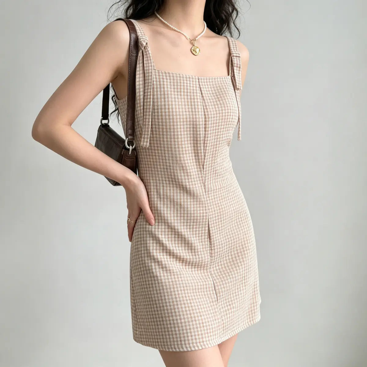 Model wearing a beige gingham mini dress with tie straps, styled with a brown leather bag and pearl necklace, side view.