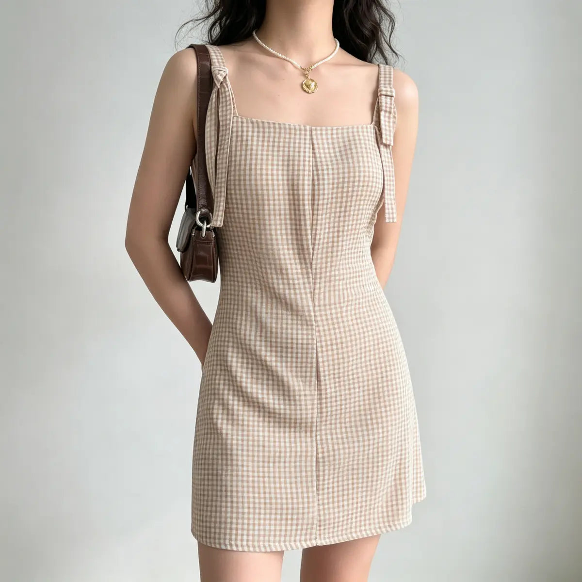 Model wearing a beige gingham mini dress with tie straps, styled with a brown leather bag and pearl necklace, three-quarter view.