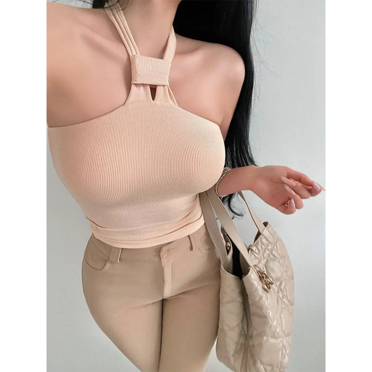  A woman wearing a beige ribbed keyhole halter top, styled with beige pants and a quilted tote bag.