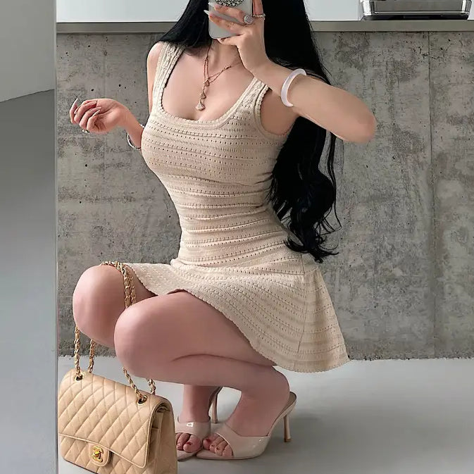 A woman squatting while wearing a beige knit mini dress with a square neckline, textured pattern, and flared skirt, styled with a beige quilted bag.