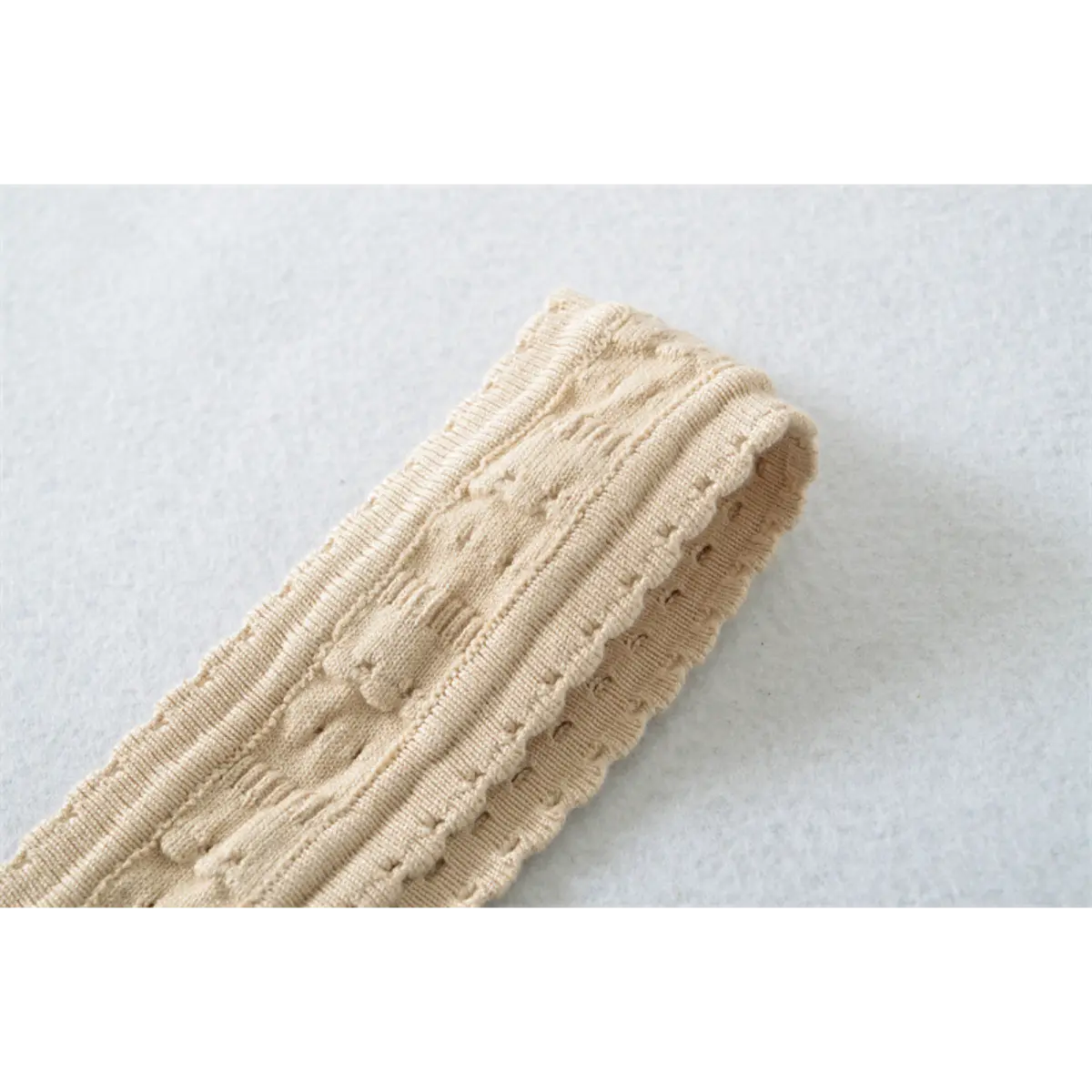 Close-up detail of the wide strap on a beige knit mini dress, showing the textured openwork pattern and scalloped edge.