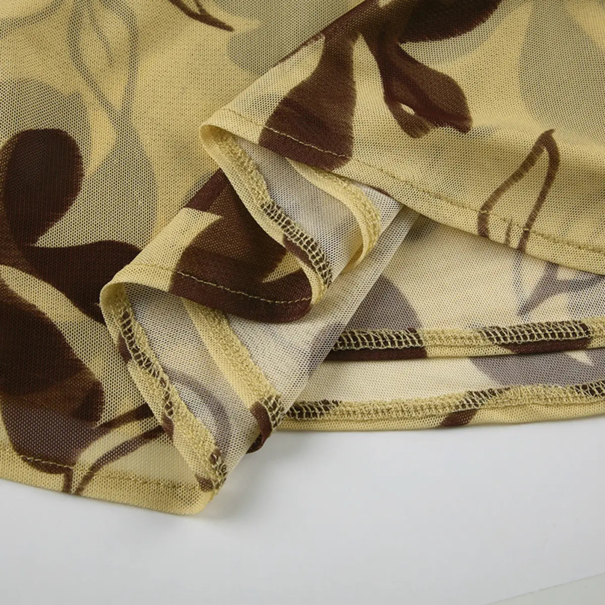 Close-up view of the hem of a beige mesh dress with brown floral print, showing the tie knot, cowl drape, and sheer fabric.
