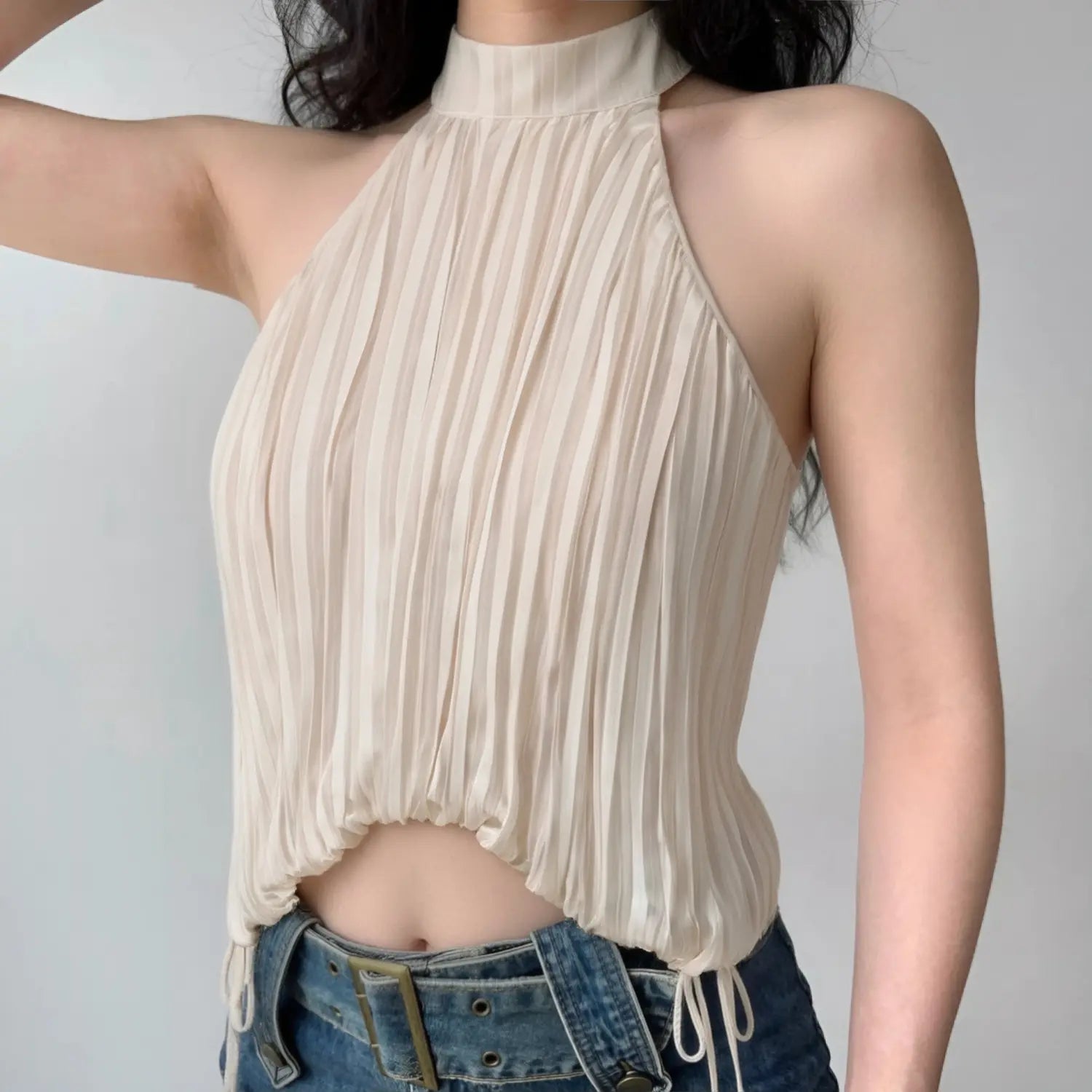 Front lifestyle shot on model of beige pleated halter cami top, folded stand collar, adjustable side ties, cropped bubble hem, sleek minimalist summer outfit