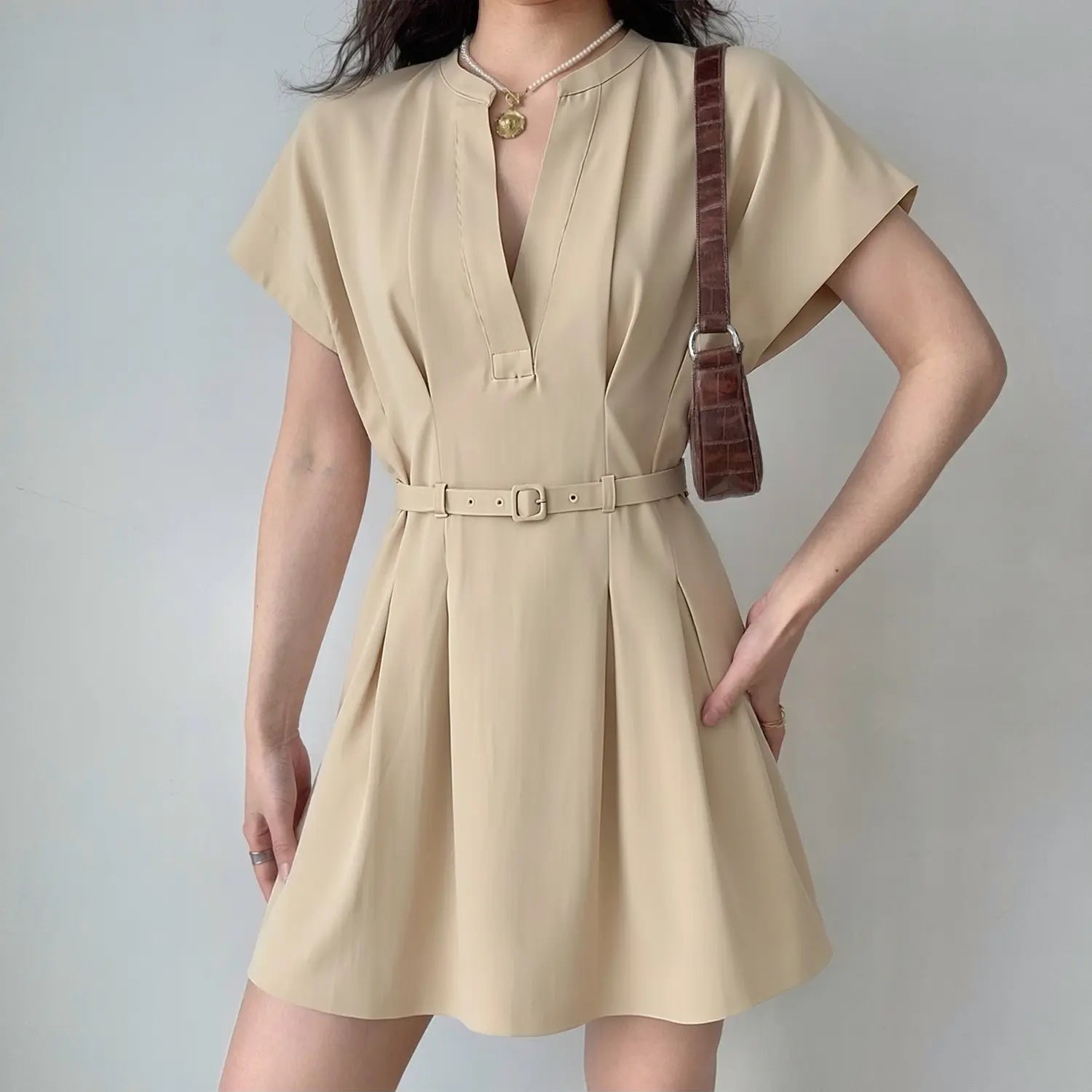 Model wearing beige v‑neck short sleeve mini dress with matching waist belt, pleated skirt, casual elegant women outfit, grey background