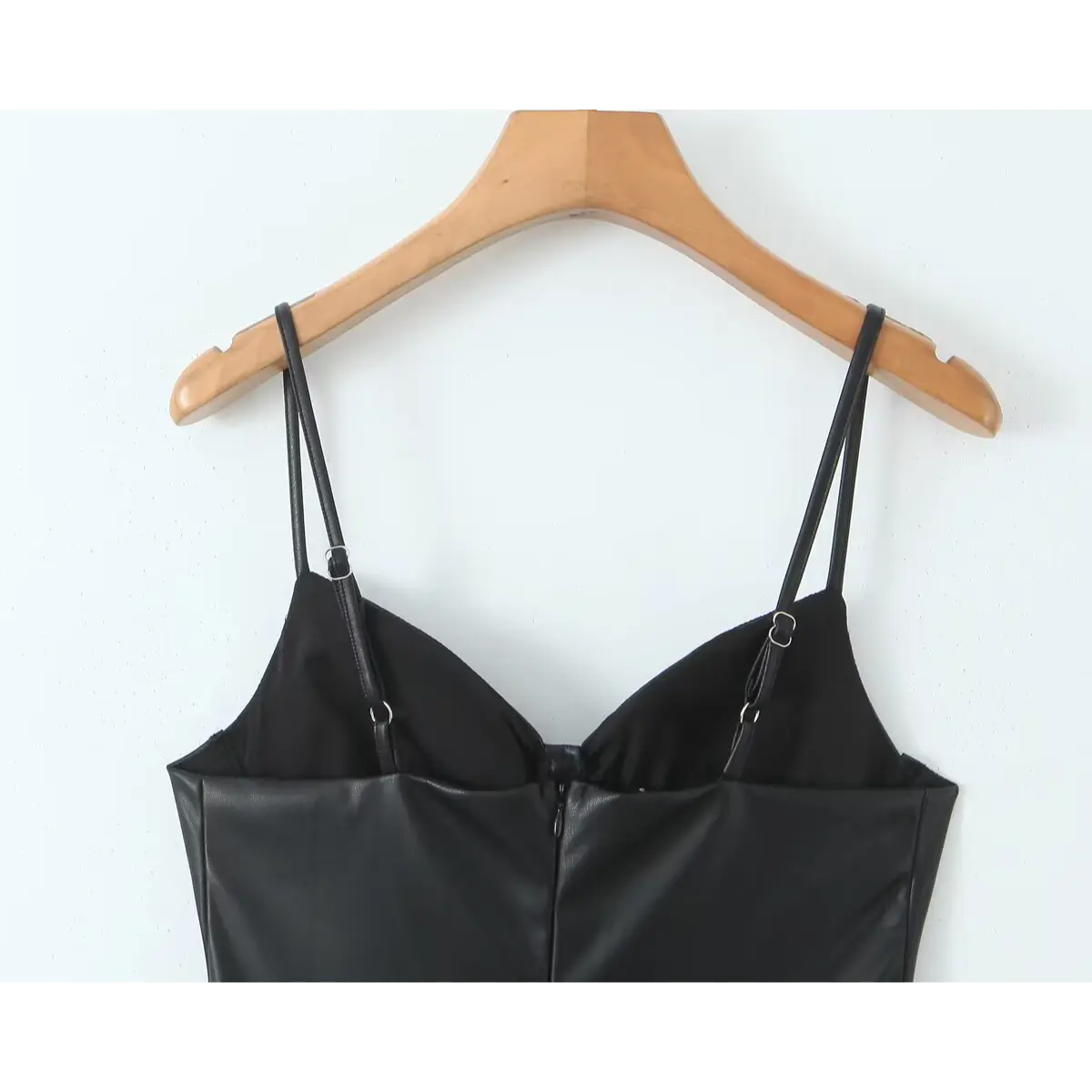 Close-up back view of a black faux-leather mini dress on a wooden hanger, showing adjustable spaghetti straps and the center-back zipper.