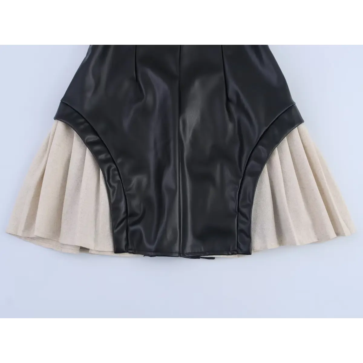 Close-up detail shot of the pleated beige skirt on a black faux-leather mini dress, showing the curved leather hem and fabric texture.