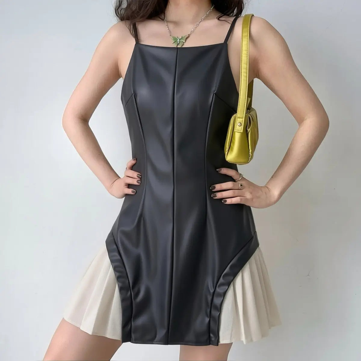 Model wearing a black faux-leather mini dress with a pleated beige skirt, styled with a yellow bag and butterfly necklace.