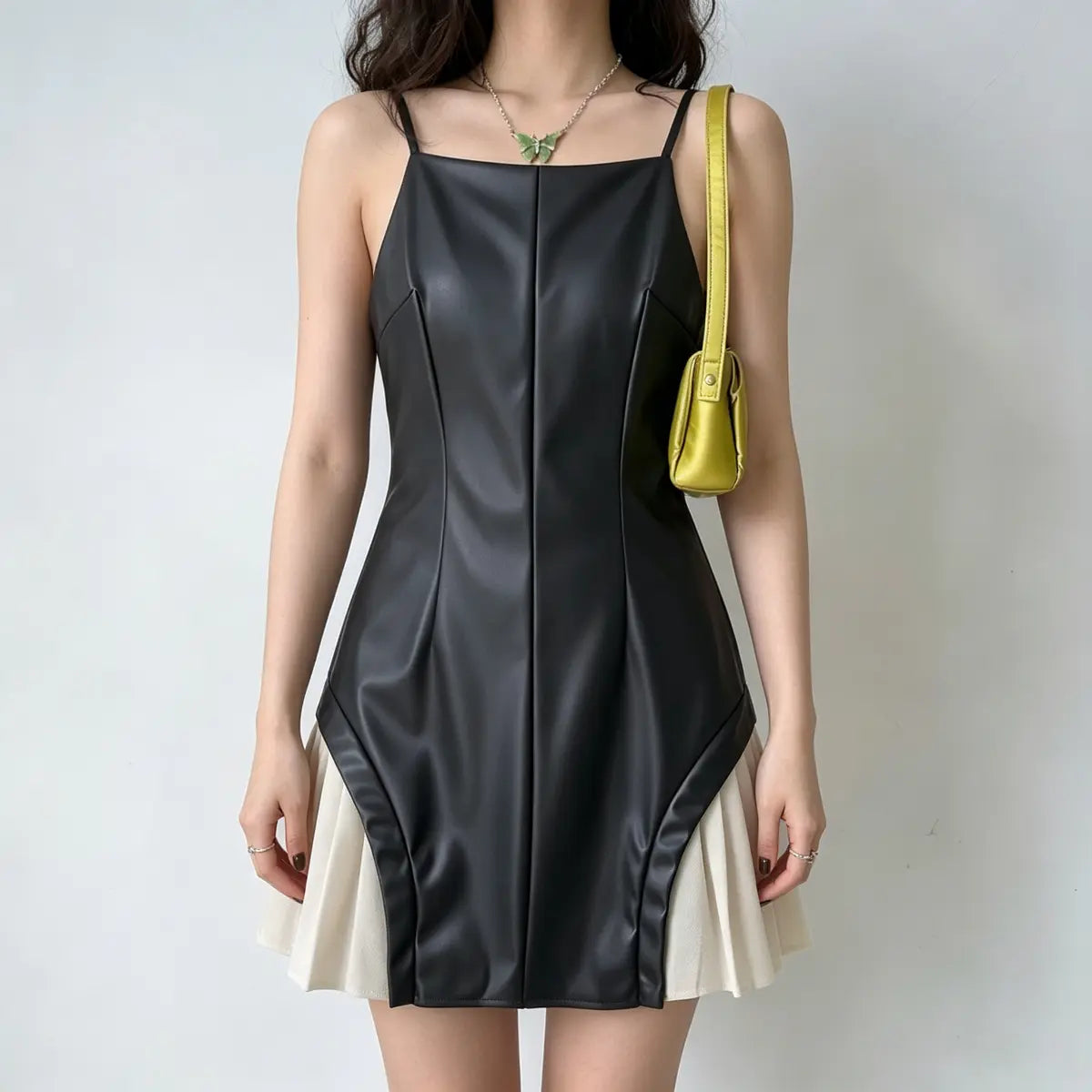 Model wearing a black faux-leather spaghetti-strap mini dress with white pleated skirt underlayer, styled with a yellow bag and green butterfly necklace, front view.