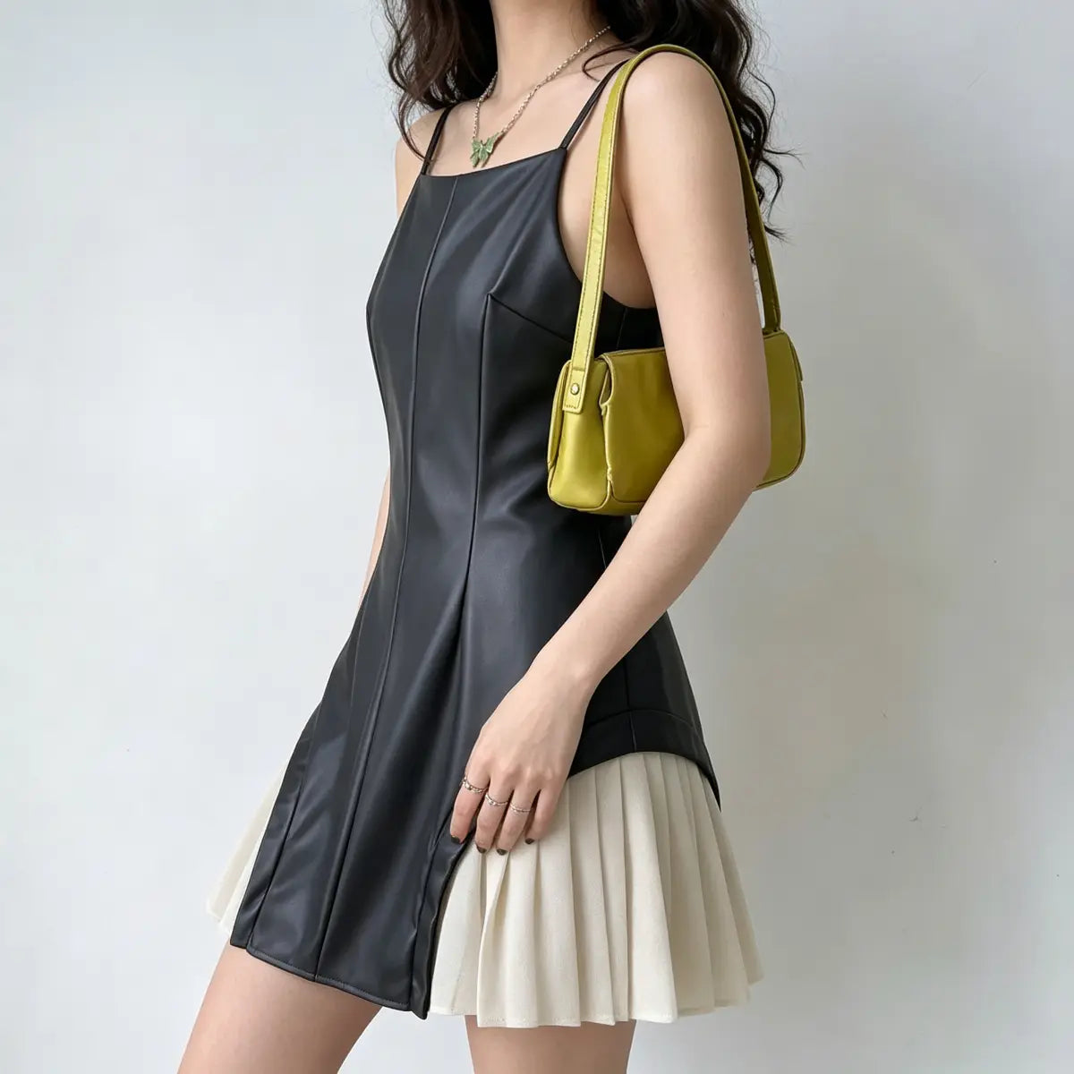 Model wearing a black faux-leather spaghetti-strap mini dress with white pleated skirt underlayer, styled with a yellow bag and green butterfly necklace, profile view.