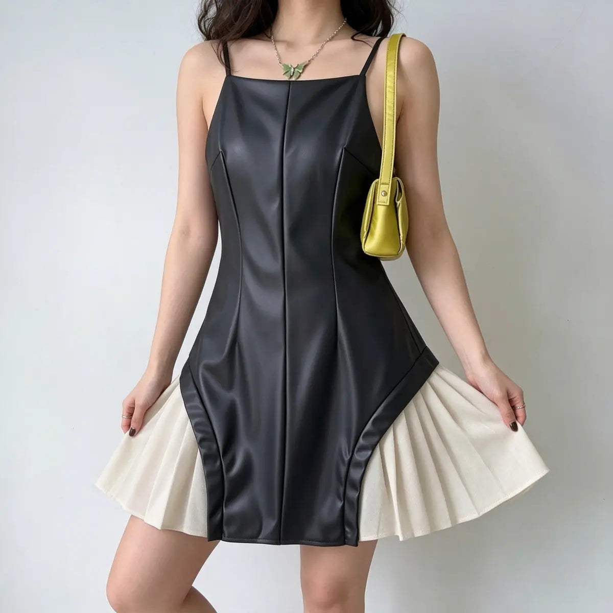 Model wearing a black faux-leather spaghetti-strap mini dress with white pleated skirt underlayer, styled with a yellow bag and green butterfly necklace, splaying the skirt to show the underlayer.