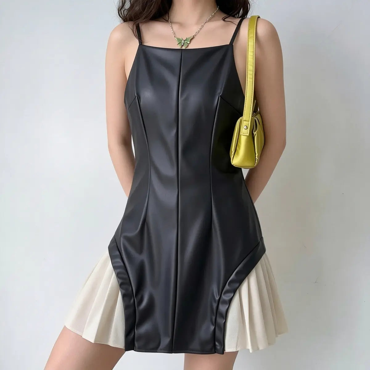 Model wearing a black faux-leather spaghetti-strap mini dress with white pleated skirt underlayer, styled with a yellow bag and green butterfly necklace, three-quarter view.