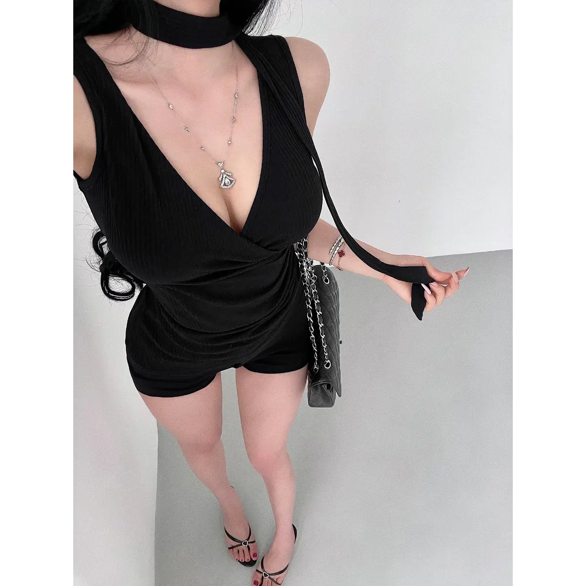 A woman wearing a black ribbed halter wrap top with a choker neckline and tie detail, styled with black shorts and accessories.