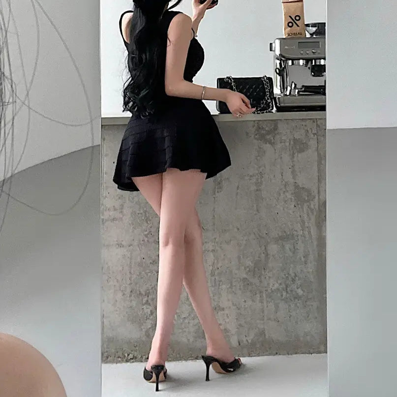 A woman wearing a black knit mini dress with a square neckline, fitted bodice, and flared skirt, taking a mirror selfie.