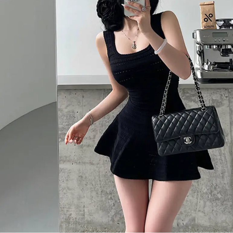 A woman wearing a black knit mini dress with a square neckline, textured pattern, and flared skirt, styled with a black quilted bag.