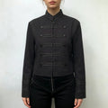 Model wearing black military inspired cropped jacket, stand collar, decorative button detailing, paired with black pants, front full torso shot